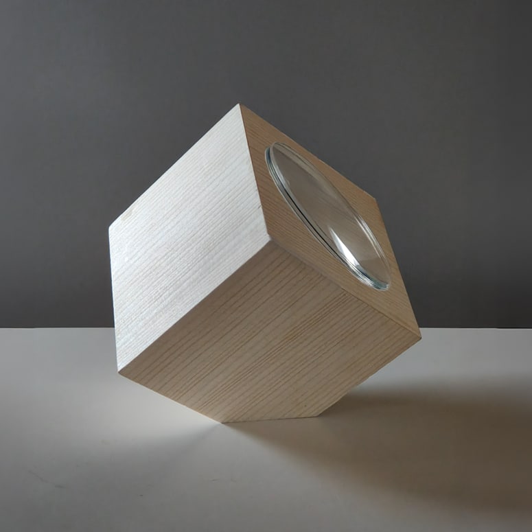 Holzwürfel aus hellem Holz mit runder Glaslinse, schräg auf einer weissen Fläche vor grauem Hintergrund