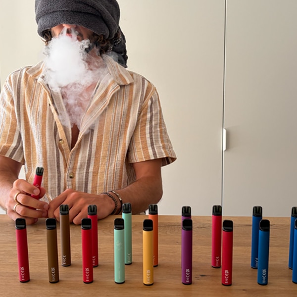 Des cigarettes électroniques jetables colorées (rouge, bleu, vert, jaune et violet) posées sur une table en bois ; une personne fume à l'arrière-plan, 20 mg